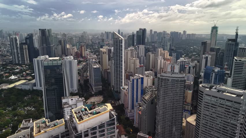 The drone approaches the Makati skyline from a mid-altitude perspective, producing a dense urban composition where multiple layers of high-rise towers fill the frame from foreground to distant horizon. Several prominent office buildings and residential towers stand close together, with darker central structures contrasted by lighter facades and reflective surfaces on surrounding buildings. The shot is wide enough to show the district context yet close enough to reveal facade lines, rooftop edges, and the varied heights that give Makati its recognizable vertical rhythm. A lower and greener urban background extends behind the main tower cluster, helping separate the business district core from the surrounding city fabric. Above the buildings, a bright overcast sky with layered cloud cover softens the scene and keeps tonal contrast moderate, which helps retain architectural detail across concrete, glass, and painted exterior surfaces. The lighting suggests late daytime without dramatic sunset color, producing a neutral palette of gray, silver, and muted blue across the architectural surfaces. Overall the image presents Makati as an active and highly concentrated metropolitan district defined by layered towers, modern development, and expansive urban depth.