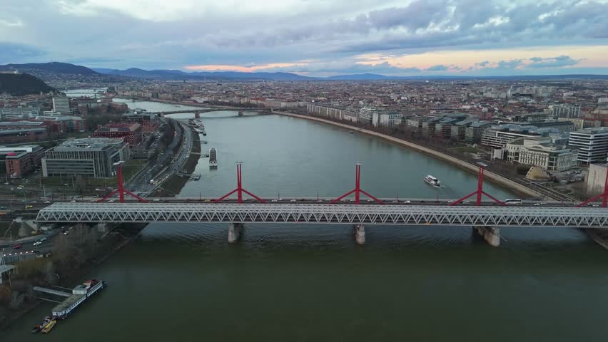 Aerial drone view of the Rákóczi Bridge spanning the Danube River in Budapest, Hungary, with urban landmarks, traffic, and the scenic sunset sky over the Buda Hills and surrounding cityscape.