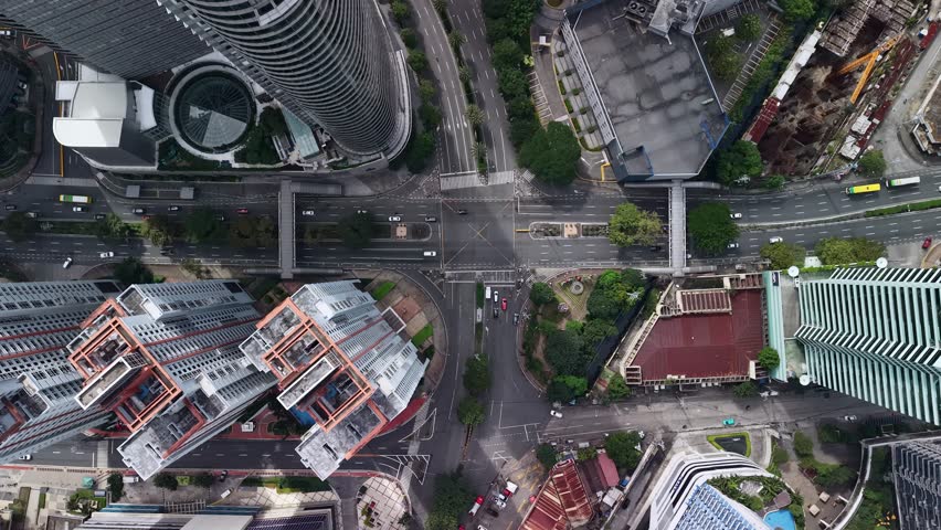 The drone hovers in a straight-down orientation above a major Makati intersection, turning the surrounding high-rise district into a precise diagram of streets, bridges, rooftops, and building footprints. A pedestrian footbridge spans across the roadway and immediately becomes the central organizing element of the frame, linking both sides of the avenue and adding a strong infrastructural feature rarely visible in standard skyline compositions. Around it, multiple traffic lanes, turning pockets, planted medians, and crossing markings reveal the complexity of circulation within the district. Curved tower edges, rectangular rooftops, and compact service areas at the corners of the frame show how the street network is compressed by dense urban development. Vehicles appear small against the wide roadway surfaces, while trees and landscaped patches soften the otherwise hard geometry of asphalt, concrete, and glass. The vertical angle emphasizes function and layout, turning the district into a readable urban map where mobility corridors and building footprints interlock. Lighting remains even and factual, with no strong directional shadows, allowing the footbridge, lane structure, and building edges to remain clear throughout the composition. The scene represents a dense and organized section of Makati where mobility infrastructure and high-rise development intersect within a compact metropolitan core.