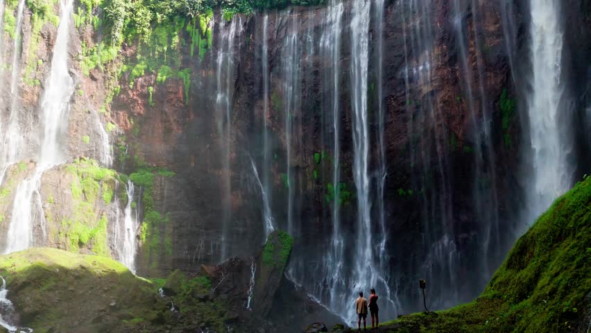 Epic 4K drone shot of Tumpak Sewu, Java, Indonesia. A couple walks toward the waterfall as the aerial camera rises, revealing towering canyon walls, flowing water, lush greenery, and misty natural bea