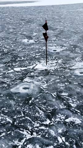 Ice fishing holes on dark frozen lake with a marker pole and snow patches, rugged winter survival scene for outdoor sport and cold-weather concepts, vertical.