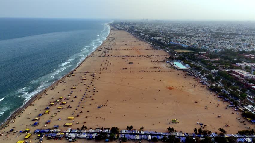 Aerial view of Marina Beach in Chennai, showcasing the vast sandy shoreline along the Bay of Bengal with waves rolling onto the coast and city neighborhoods stretching along the eastern edge.