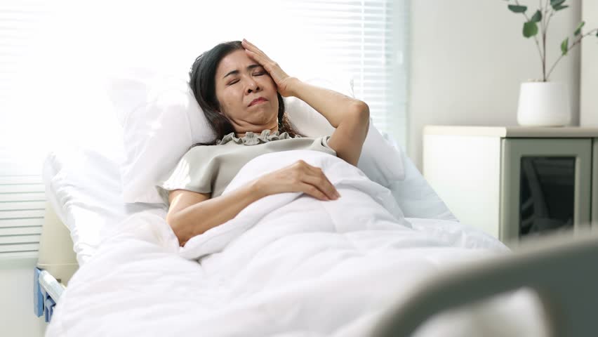 Hospitalized elderly Asian woman experiencing intense headache while resting in hospital bed, discomfort and physical pain from health condition.