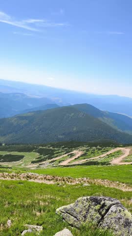 Expansive view from a mountain summit, showing lush green slopes, rocky terrain, and a winding trail under a clear blue summer sky in Demanovska Dolina, Slovakia, Chopok