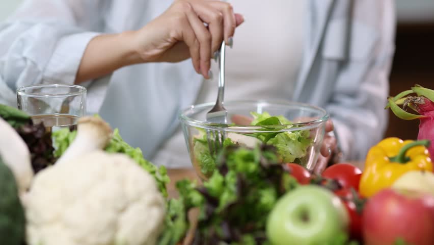 Young Asian woman in casual clothes is sitting at a wooden table, eating a healthy salad from a glass bowl, surrounded by a variety of fresh fruits and vegetables.