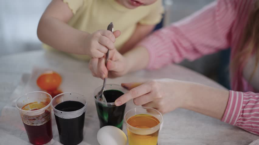 Little girl holds egg, dips it slowly in colorful dye. Mother guides her gently, smiling warmly. Soft light shows cozy, joyful Easter activity. Perfect for family, holiday, or craft projects
