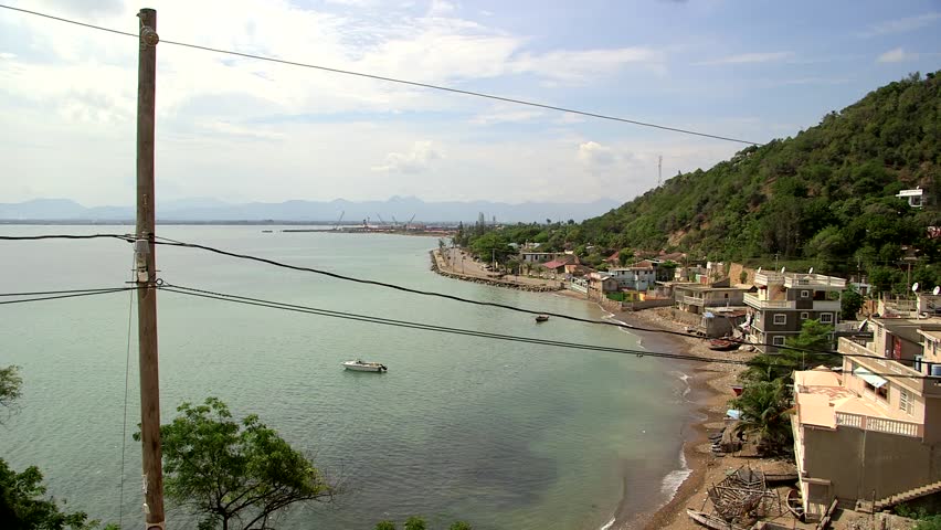 Panoramic view of Cap-Haitien coastline in Haiti, with colorful hillside buildings along turquoise bay, small boat on water, distant mountains, power lines, and lush green hills under partly cloudy sk