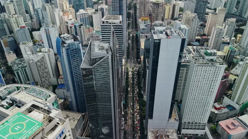 The drone holds a wide elevated view over the Makati financial district, centering a long urban avenue while surrounding office towers and high-rises extend outward to fill most of the frame. Compared with tighter city canyon shots, this composition gives more breathing room around the vertical core and shows how the business district connects to lower neighborhoods and greener areas beyond the dense cluster. The central corridor acts as the main organizing line, with moving vehicles visible below and repeated building facades stepping backward into the distance. Architectural variety is clear, including glass curtain walls, white office blocks, blue-toned towers, rooftop mechanical structures, and cylindrical or rectangular forms that break up the skyline. Layered clouds cover the sky and keep the light soft, which helps preserve detail in bright rooftops and reflective surfaces without harsh contrast. If readable logos remain visible in the full clip this shot should be treated as editorial, but visually it functions as a broad establishing aerial of Makati