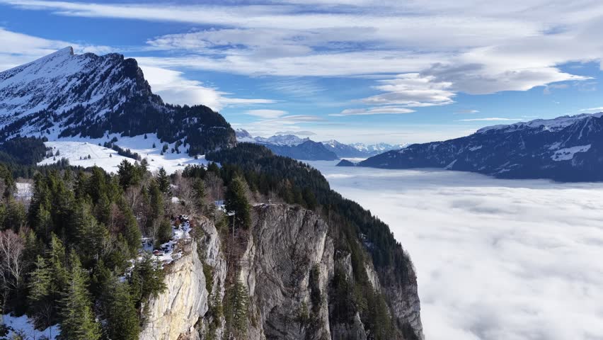 Snow covered mountain cliff above dense forest and a valley filled with low clouds and distant peaks