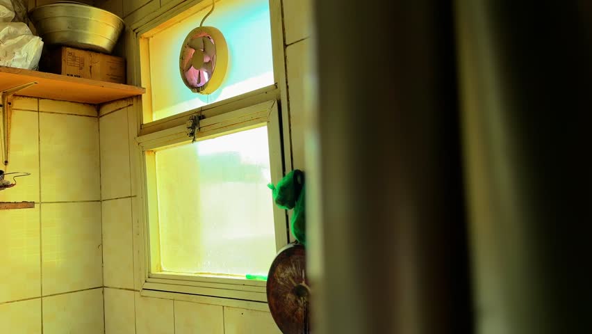 Warm sunlight entering through a frosted kitchen window with a small ventilation fan, rustic shelves, and cookware in a modest home interior, capturing everyday domestic life and natural indoor lighti