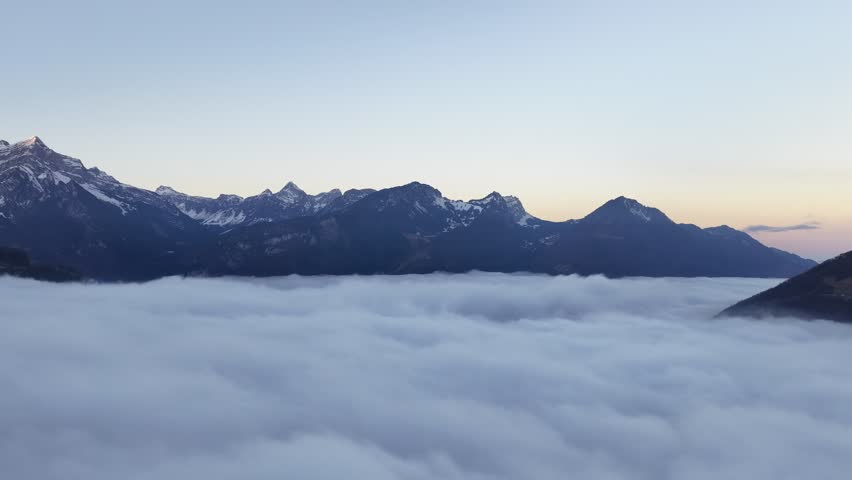 Aerial view above sea of clouds with alpine mountains silhouettes during sunrise in dramatic landscape.