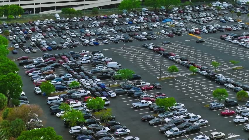 Aerial view shows a large parking lot in Miami filled with cars. Nearby streets have some traffic, while trees line the edges, adding some greenery to the urban setting.