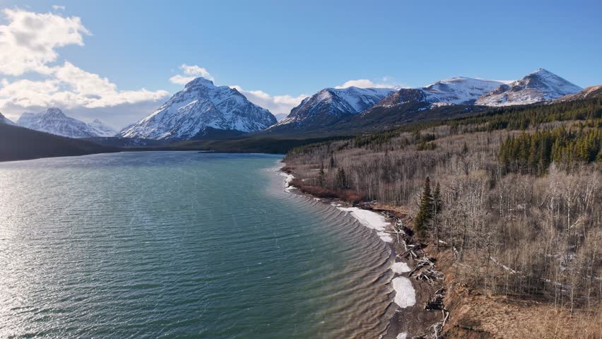 Aerial view of a pristine lake and shoreline with snow capped mountains and coniferous forests in Montana.