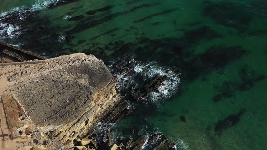 Aerial drone shot of rugged rocky shoreline with waves rolling over reefs, showing clear green coastal water and seaside landscape during sunny day