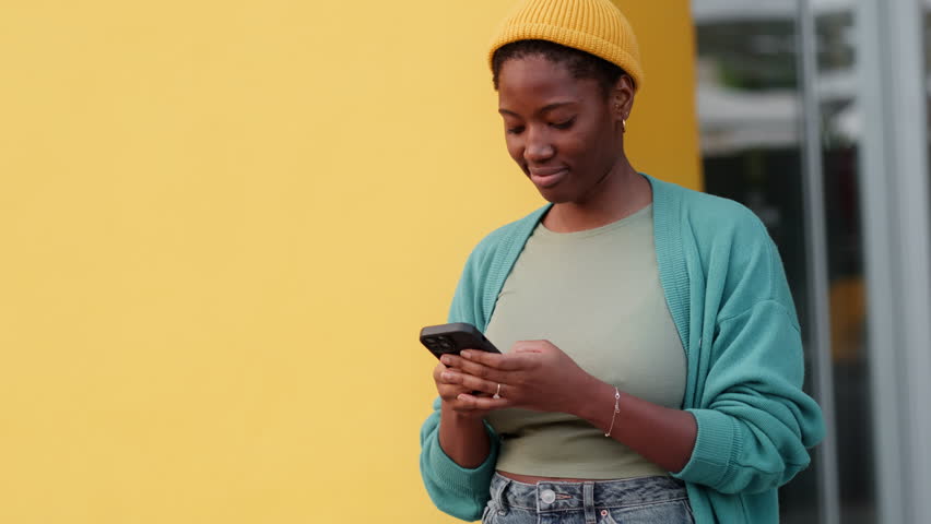 Smiling young Black woman using smartphone against yellow wall. Happy female in yellow cap text messaging on phone while looking around on city street