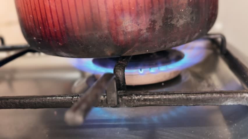 Close-up of a metal cooking pot heating on a gas stove burner with visible blue flame in a home kitchen, capturing everyday cooking activity and traditional stovetop food preparation.