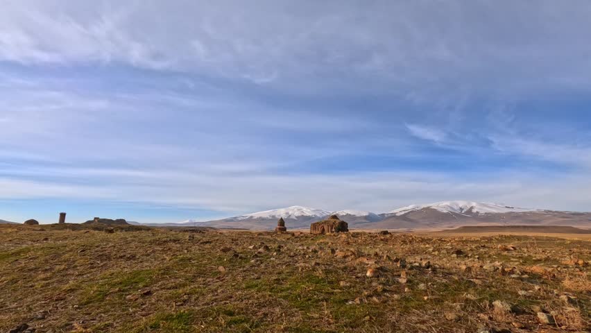 Wide view of Abughamrents Church and Tigran Honents Church in Ani, Kars, Turkey. Historic Armenian medieval structures set in vast steppe landscape with snowy mountains under dramatic sky.