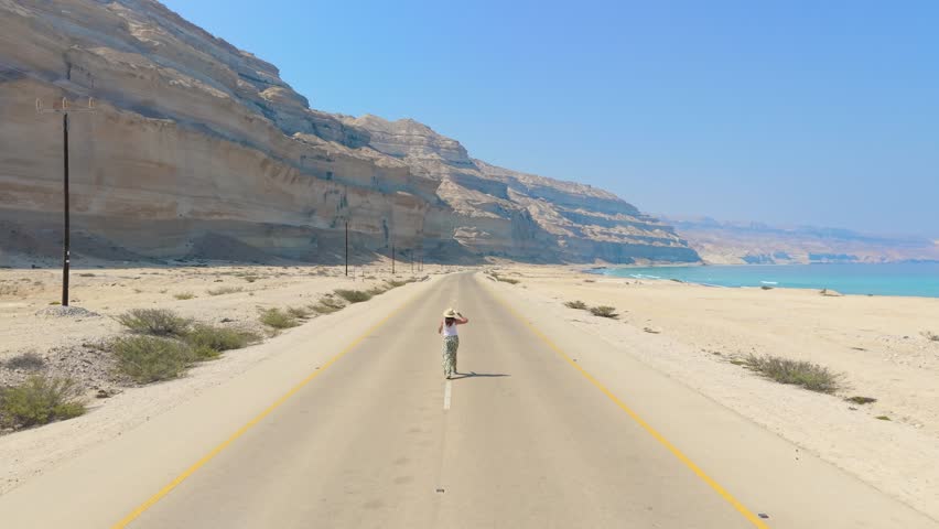 Aerial view of a young woman walking along an empty coastal road between high desert cliffs and the turquoise ocean in Salalah, Oman.