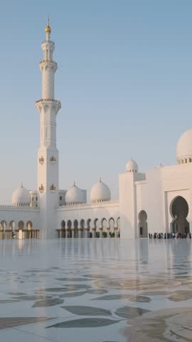 Grand courtyard and entrance of Sheikh Zayed Grand Mosque with white domes, minaret and reflections on marble floor under clear blue sky in Abu Dhabi, United Arab Emirates. Arabic architecture