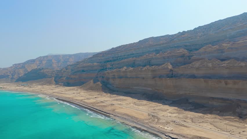 Aerial view of turquoise water along a sandy coastline with tall sedimentary cliffs and a desert road in Salalah, Oman.