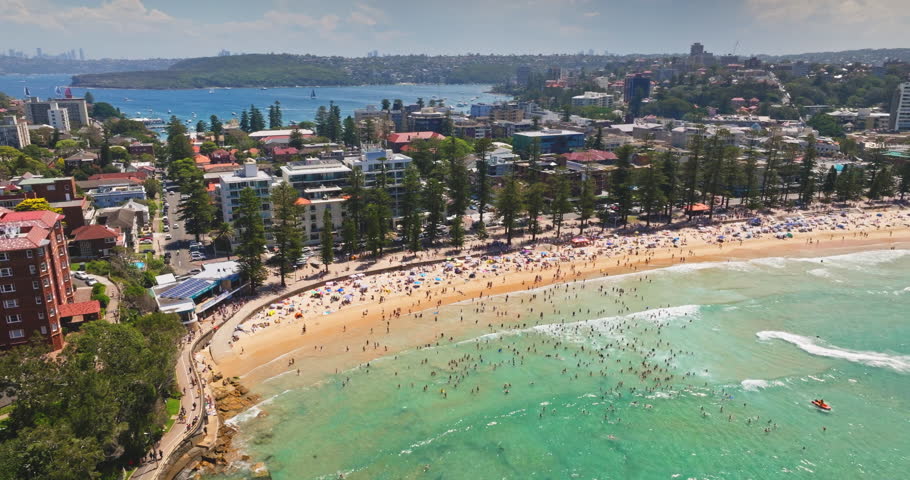 Crowds people on Manly Beach enjoying a sunny summer day, turquoise waters and sandy shore framed by coastal buildings and lush greenery in an aerial panoramic view. Travel to Australia