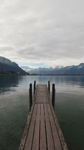 Person walking on a wooden pier toward the Alps on Lake Geneva near Montreux, Switzerland. Vertical video format ideal for social media, mobile content and modern digital advertising. Scenic European lakeside landscape.