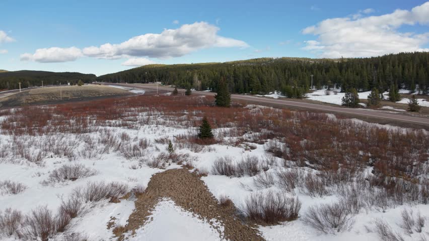 Aerial view over a snowy marsh and shrubs with small evergreen trees, winding road, and distant snow capped mountains under partly cloudy sky.