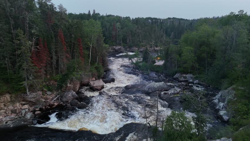 Turtle Falls on the Manigotagan River in Manitoba flowing over Canadian Shield granite. The aerial camera flies downstream along rapids past a riverside tent and toward calmer flat water.
