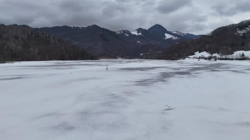 Aerial view of a frozen lake surrounded by mountains and forests under a cloudy sky