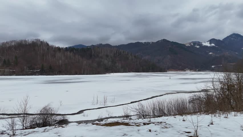 Drone shot flying over a frozen mountain lake surrounded by snowy peaks and forests