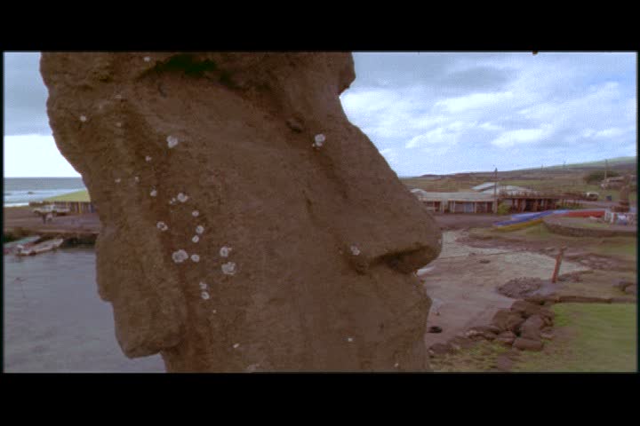 EASTER ISLAND, CHILE - FEBRUARY 15, 2000: CU moai, camera cranes down to show WS of coastal village and men paddling out in outrigger canoe.