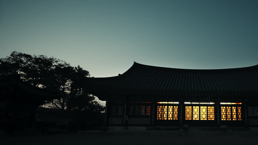 Cinematic wide shot of traditional Korean temple at dusk with glowing windows illuminating courtyard under deep blue twilight sky