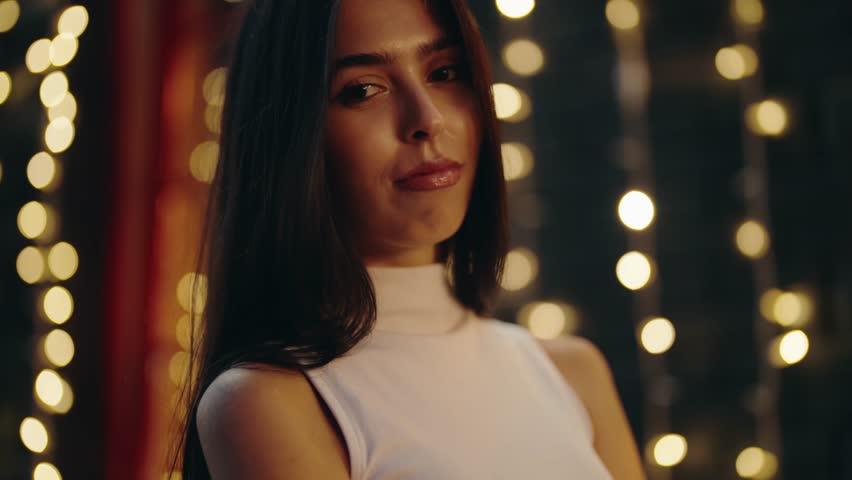 Charming young woman with long dark hair looking seductively at the camera and smiling gently while posing at night with warm, golden bokeh lights creating a magical atmosphere