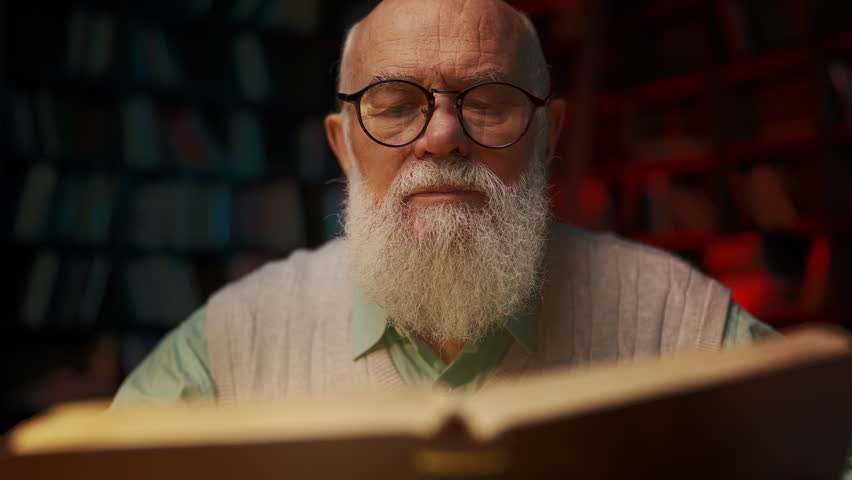 Focused elderly male scientist reading book late at night, absorbing knowledge
