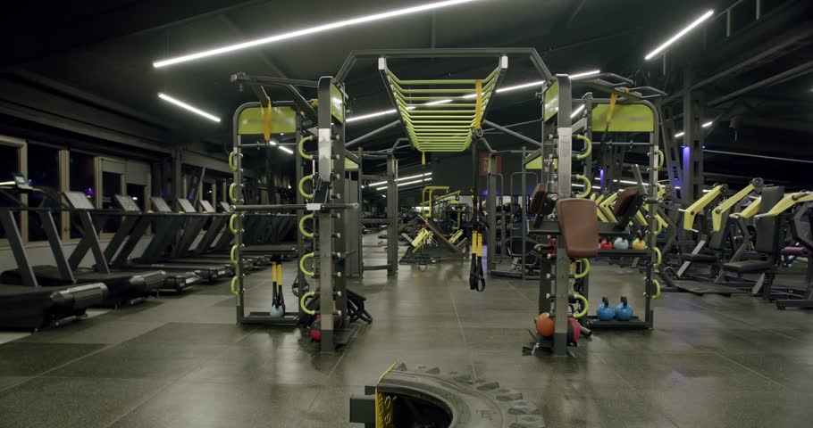 Empty modern gym interior featuring various workout equipment, including treadmills, a functional training rig, and kettlebells, illuminated by contemporary ceiling lights for a sleek look