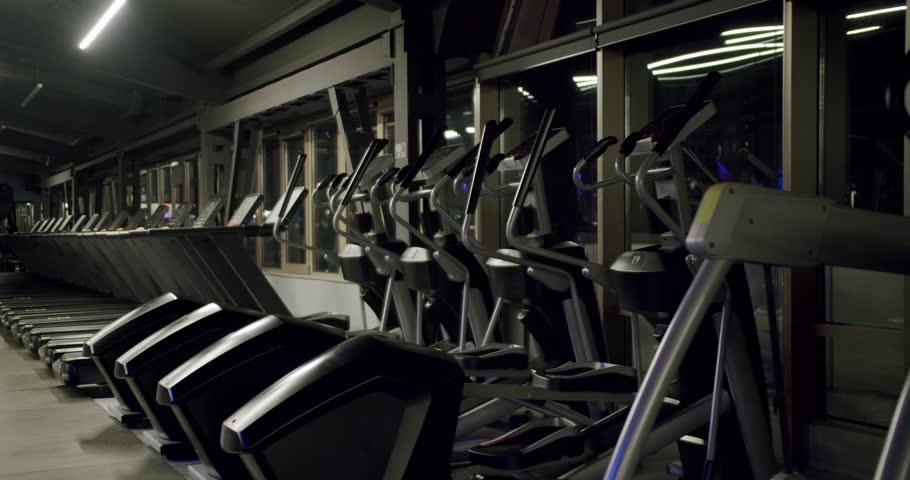 Dark interior of an empty fitness club at night with rows of modern treadmills and elliptical cross trainers standing ready for a workout, reflecting the city lights from the large windows