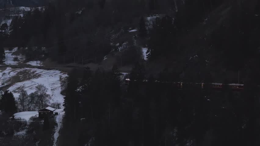Aerial view shows the Glacier Express crossing Landwasser Viaduct near Filisur in Graubunden, entering a dark gorge, with snow dusted slopes, chalets, and a farm road.