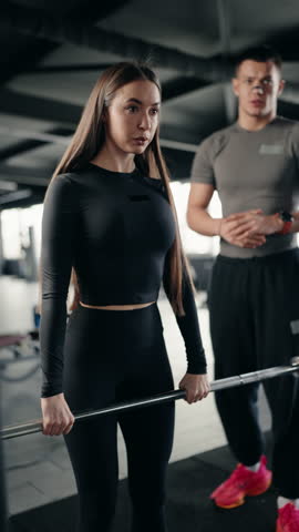 A young woman performs deadlifts with a barbell in a gym, guided by her personal trainer. She focuses on proper form and technique as she lifts. vertical