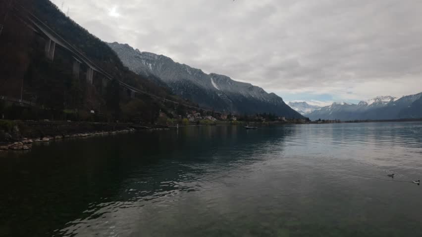 Scenic view of Lake Geneva with Alpine mountains near Montreux, Switzerland. A train passes along the lakeside while calm water reflects the cloudy sky. Peaceful Swiss landscape with transport element and copy space.
