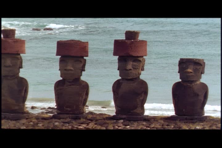 MS camera pans left over row of moai statues with pukao or topknots on Easter Island, Chile. Blue waves wash up onto beach in background.
