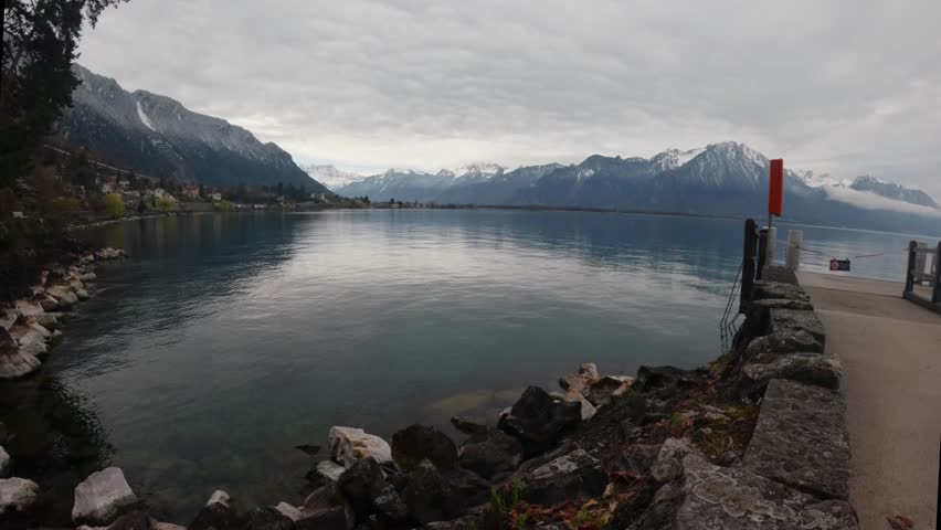 Scenic view of Lake Geneva near Montreux, Switzerland, featuring a lakeshore walkway with rocks and a red navigation marker. Calm water and Alpine mountains under a cloudy sky create a peaceful Swiss landscape while a distant train moves along the lakeside railway.