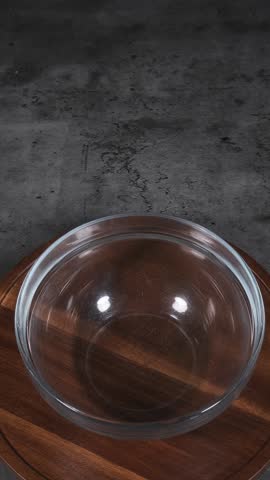 High angle vertical 1080p footage of vibrant blue bubble gum flavoured liquid chocolate pouring from off-camera into a clear glass bowl. Cinematic lighting on a neutral grey table background highlights the smooth, glossy flow.