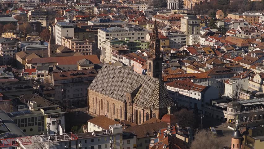 Aerial pans over Bolzano, Italy, centering on Duomo di Bolzano and Piazza Walther. Diamond roof and tall bell tower rise above red tiles in warm late day light.