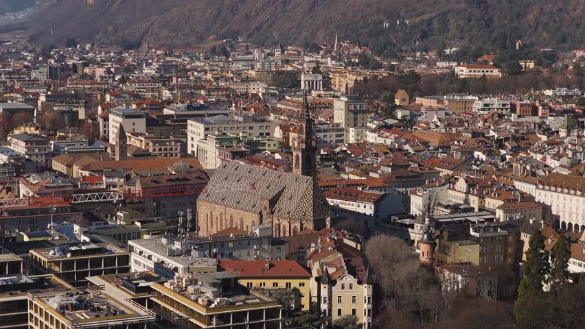 Aerial view of Bolzano, Italy, with a Gothic cathedral, diamond tiled roof, tall spire, terracotta rooftops, a red crane, and Alpine slopes in crisp sunlit tones.