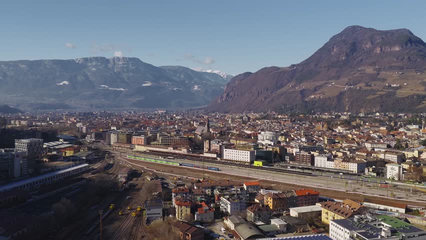 Aerial winter panorama of Bolzano, Italy, with the central railway station, active rail yard, green regional trains, historic town center, and terraced Alpine slopes.