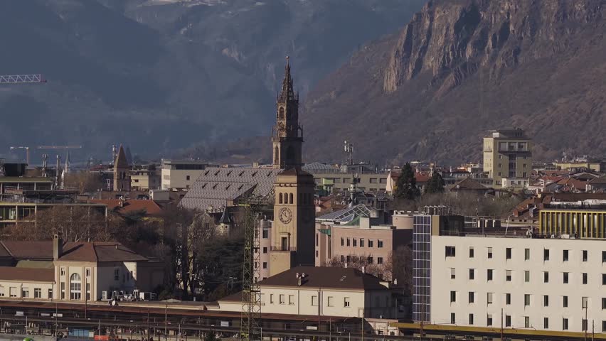 Aerial view of Bolzano, Italy, with Duomo spire and diamond roof. Camera pans to alpine cliffs and Adige valley. Warm low light, wide frame, bare trees, cranes.