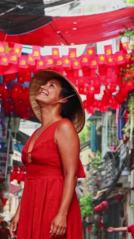 Happy tourist woman wearing a traditional vietnamese conical hat and a red dress smiling in a decorated street in Hanoi. Celebration of the Communist Party of Vietnam with red flags
