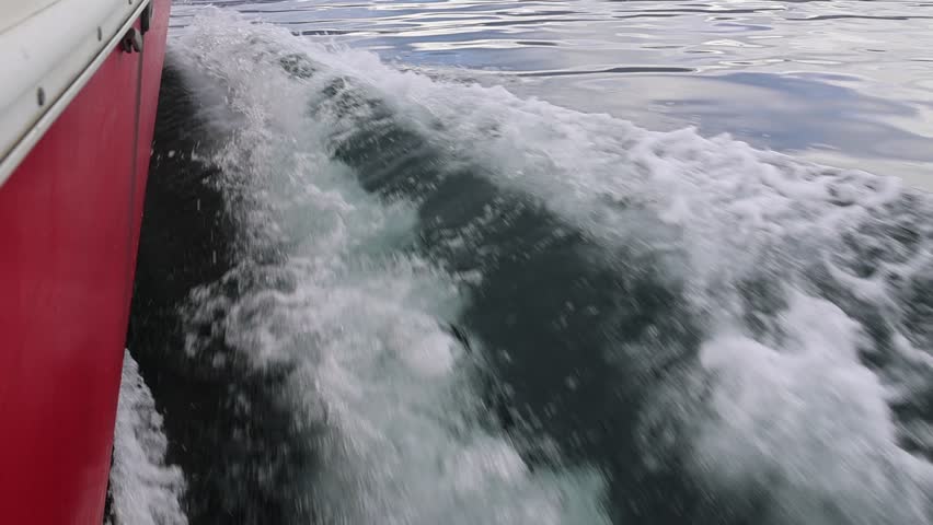The red hull of a small boat is visible on the left side of the video, while on the right side, water forms ripples and foam due to the boat's movement. The video is in slow motion and at a close-up angle.