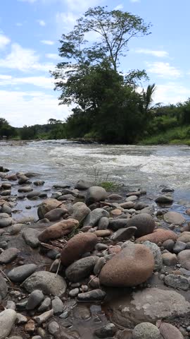 Rushing River Stream with Smooth Stones and Trees 

A beautiful landscape featuring a fast-flowing river with white water rapids, surrounded by smooth river stones and lush green trees under a blue sky.
