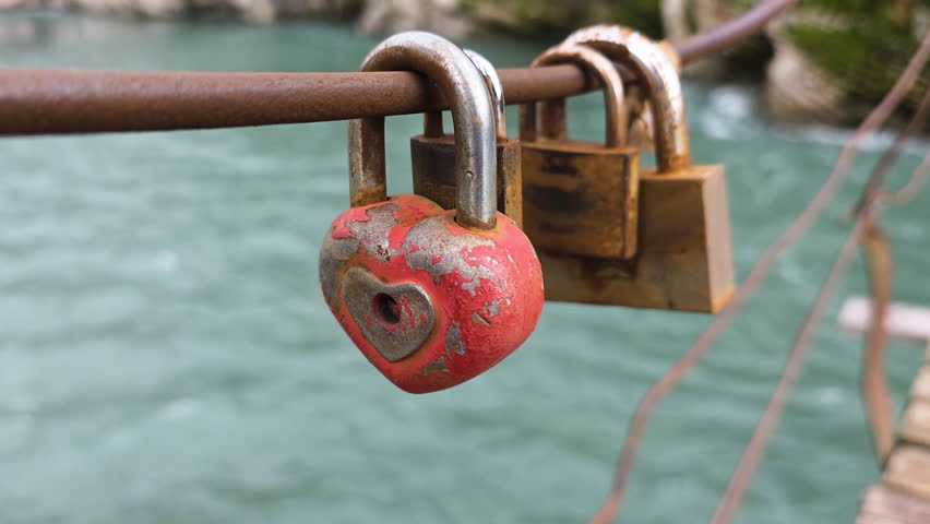 Heart-shaped love lock swaying on a rusty bridge rail above a flowing river, symbolizing eternal love and commitment